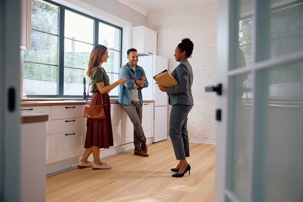 A smiling couple examines a home’s kitchen while an inspector points to features on a notepad.